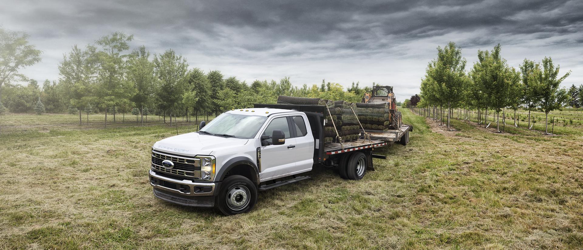 A White 2026 Ford Super Duty® Chassis Cab hauling something and towing a trailer with a wheeled skid steer