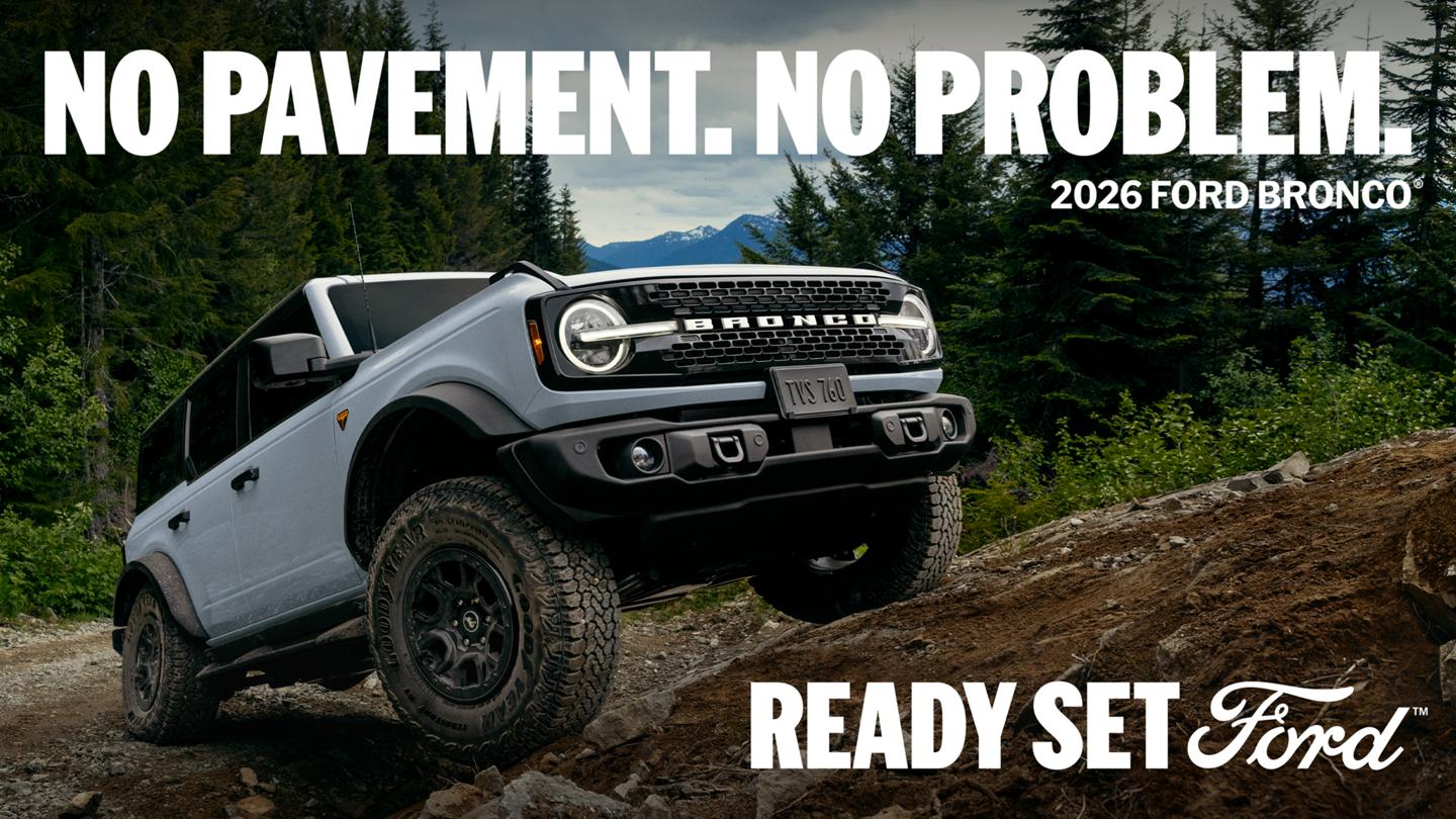 A front three-quarter view of a grey, four-door 2026 Ford Bronco climbing a steep, rugged dirt hill, set against a pine forest background. Bold text reads "NO PAVEMENT. NO PROBLEM. 2026 FORD BRONCO" at the top and "READY SET Ford" at the bottom right.
