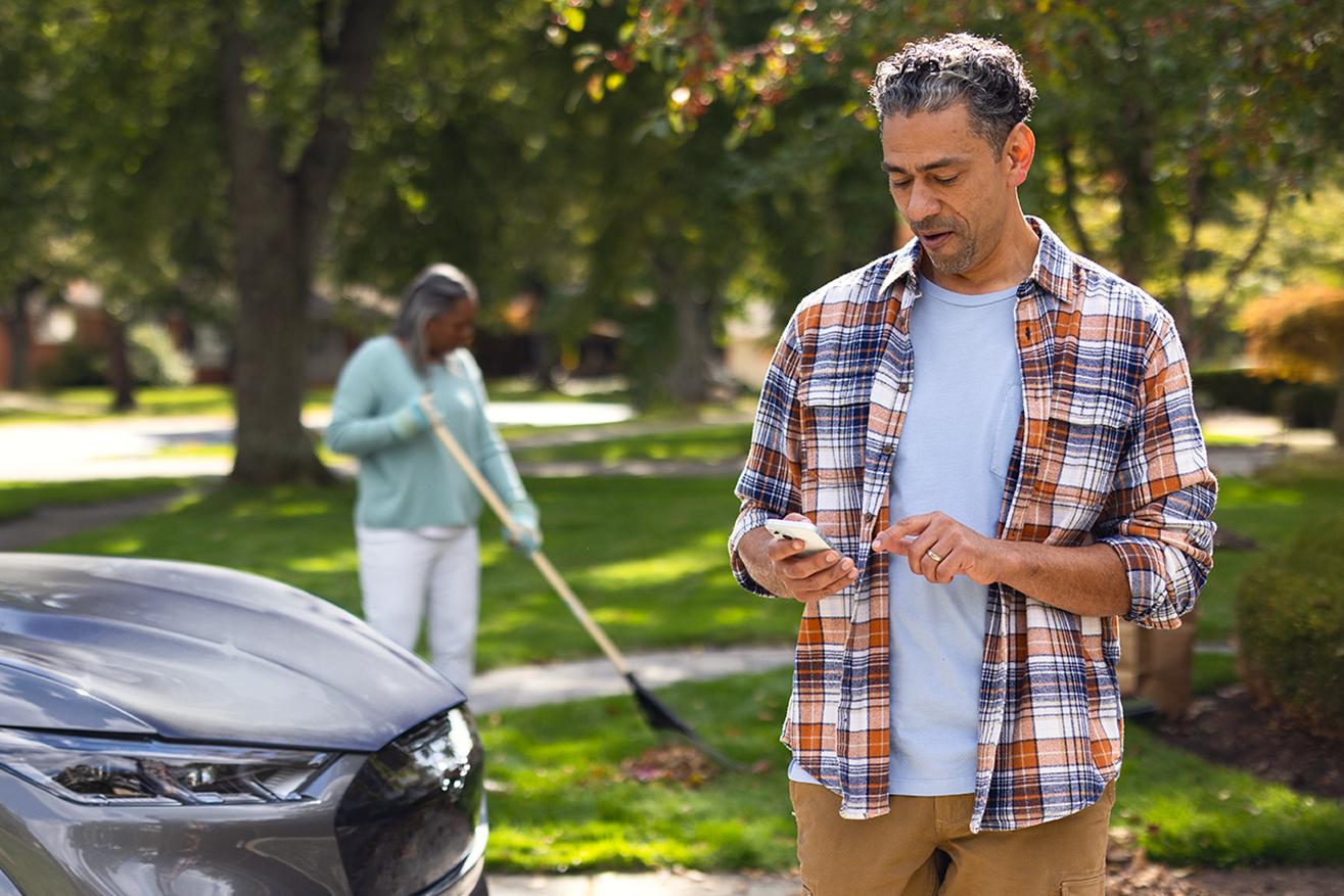 Two adults are outside near their Mustang Mach-E SUV. One rakes leaves, while the other checks their phone.