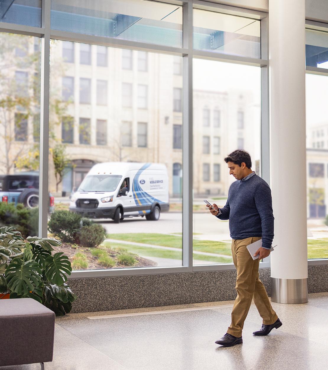 A person walks past a window inside a building while looking at their phone. Outside, a Mobile Service van is arriving.