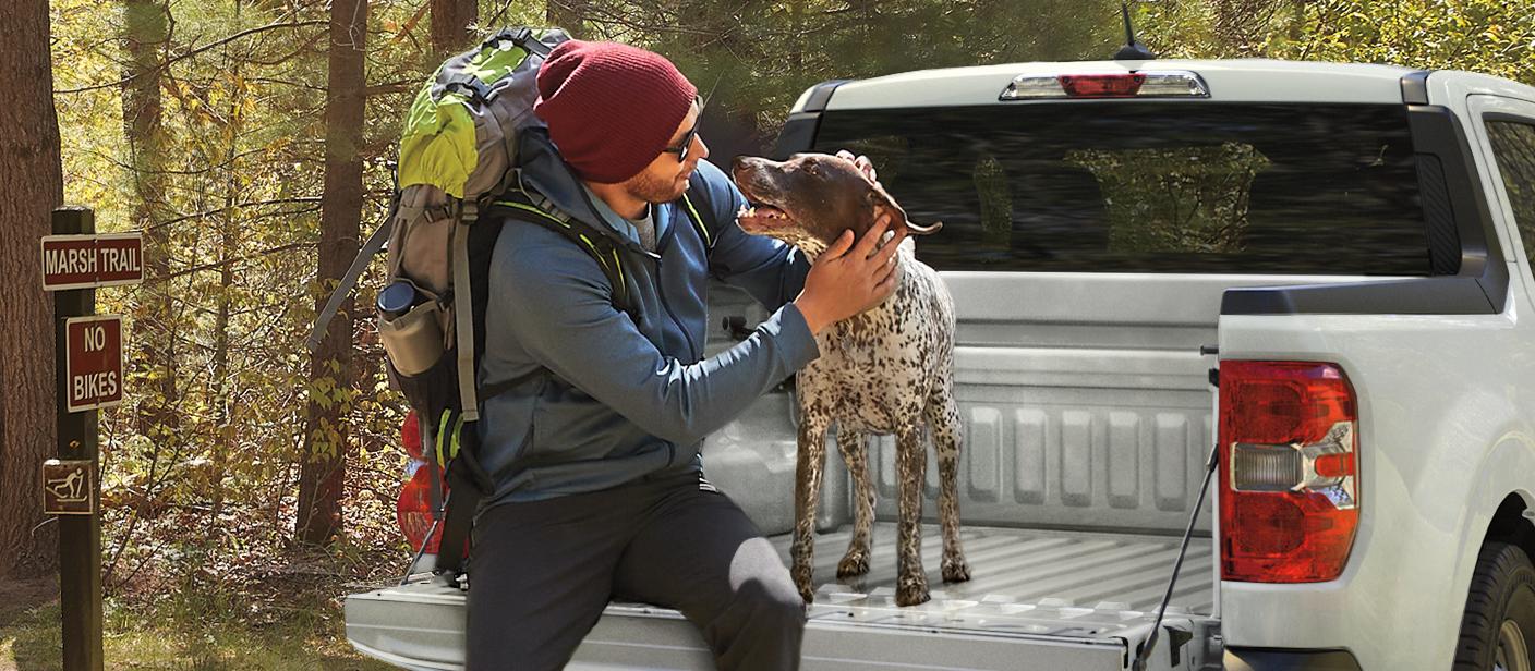 Person and a dog sit on the tailgate of a Ford Maverick parked next to a trail.