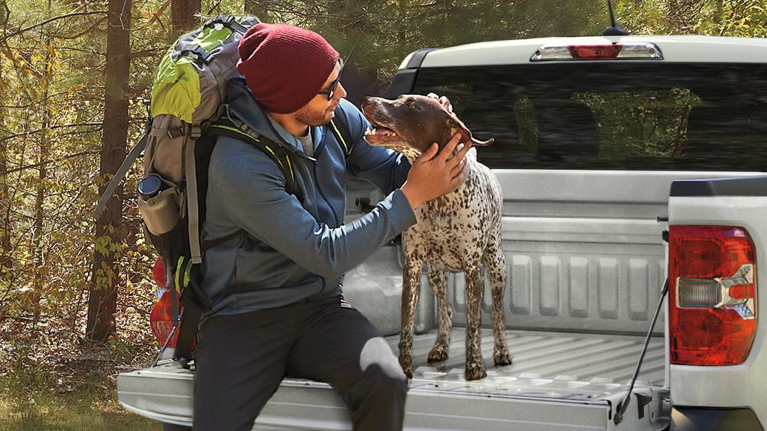 Person and a dog sit on the tailgate of a Ford Maverick parked next to a trail.