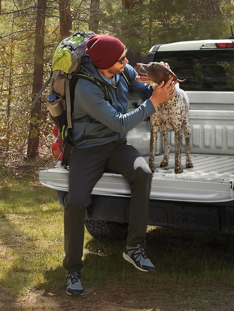 Person and a dog sit on the tailgate of a Ford Maverick parked next to a trail.