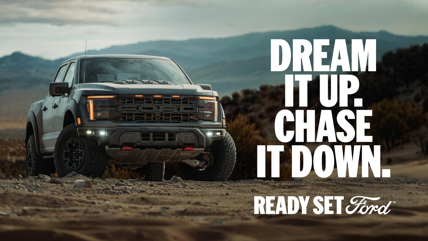 Three-quarter front view of a Ford F-150 Raptor parked on dirt with mountains in the background. "Dream it up. Chase it down. Ready Set Ford."