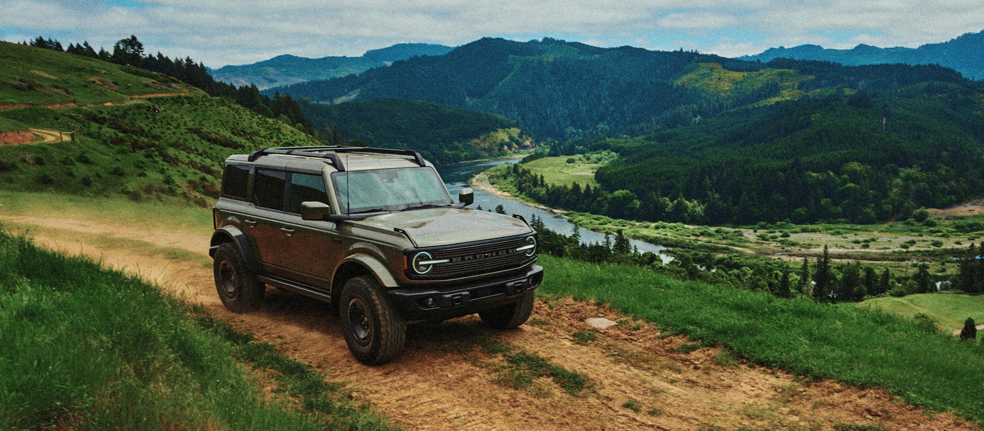 A 2027 Ford Bronco® Filson SUV being driven on a dirt track along the crest of a hill, overlooking a river valley
