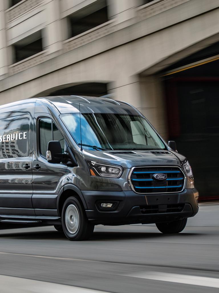 A dark grey 2027 Ford E-Transit™ Cargo Van with blue grille accents driving on a city street alongside a modern building