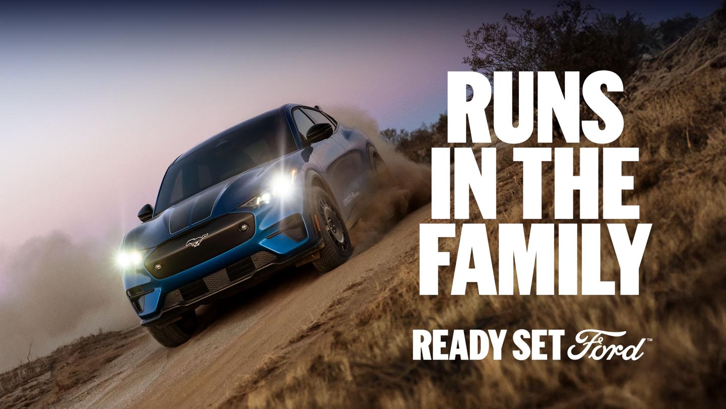 Three-quarter front view of a blue Ford Mustang Mach-E electric SUV driving on a dirt road at an angle. "Runs in the family. Ready Set Ford."