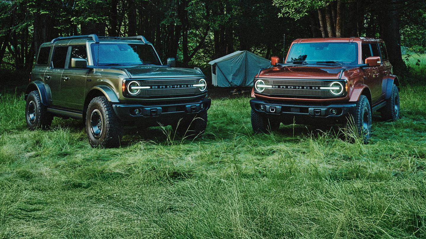 Two Ford Bronco SUVs parked side-by-side in a three-quarter front view, at a forested campsite. A dark green Bronco is on the left, and a copper-red Bronco is on the right.