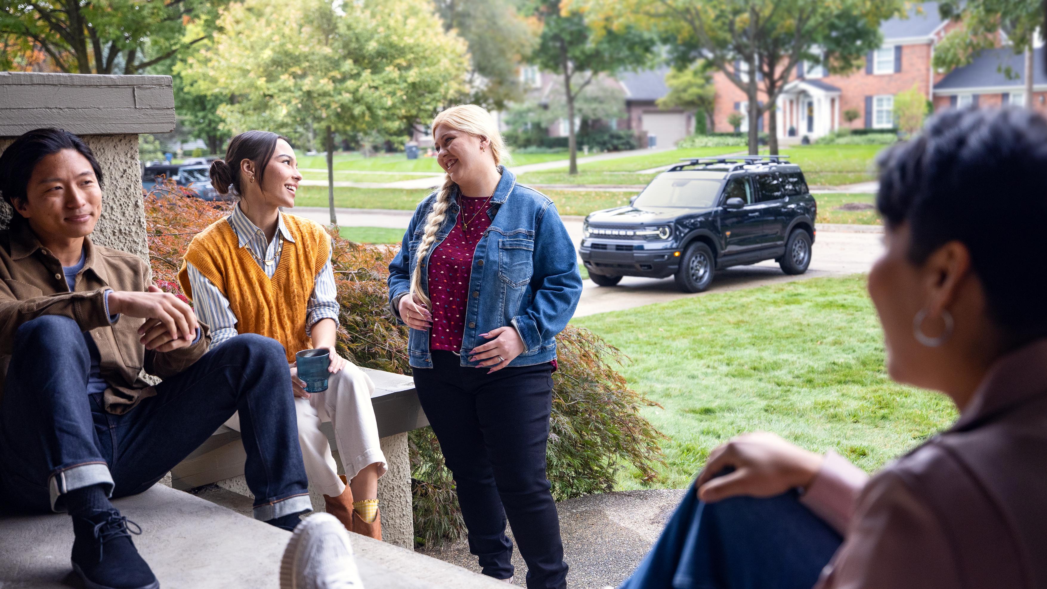 A group of people gather on the front porch of a home with a Ford Bronco parked in the driveway.