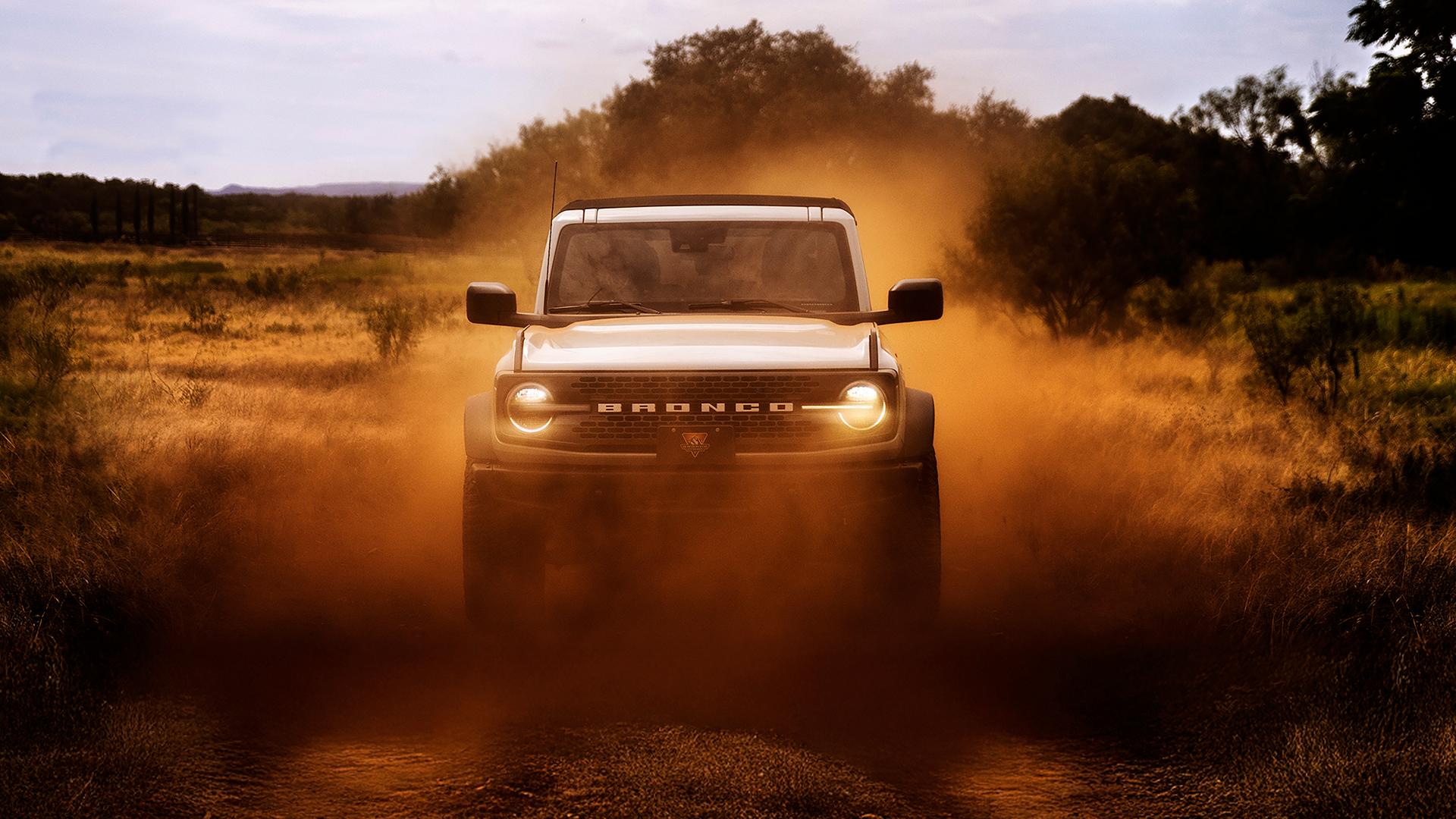 The front of a 2026 Ford Bronco® SUV, its headlights shining through a cloud of red dust