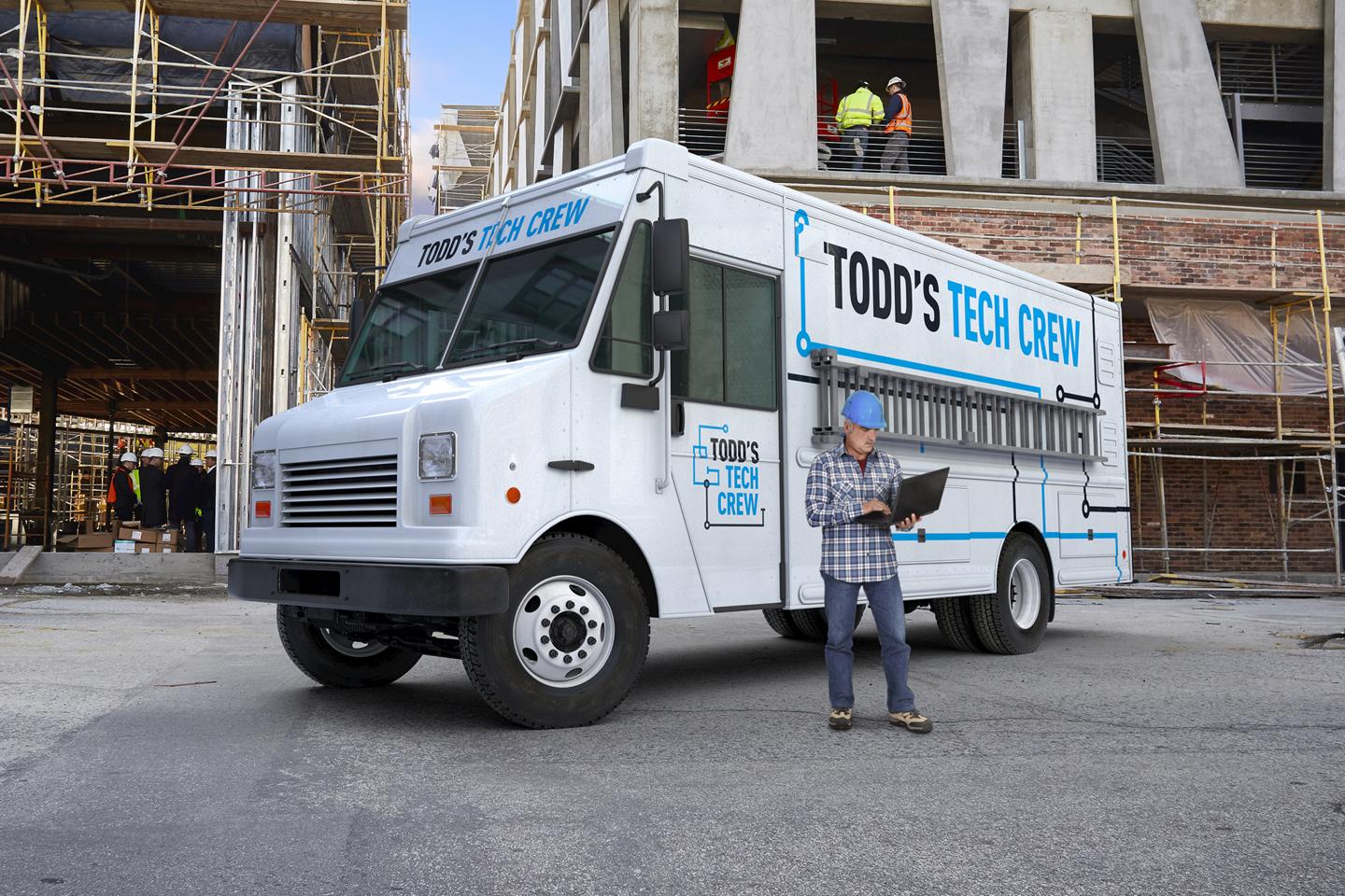 A man stands holding a laptop in front of a 2025 Ford F-59 Stripped Chassis that is parked in front of a building under construction