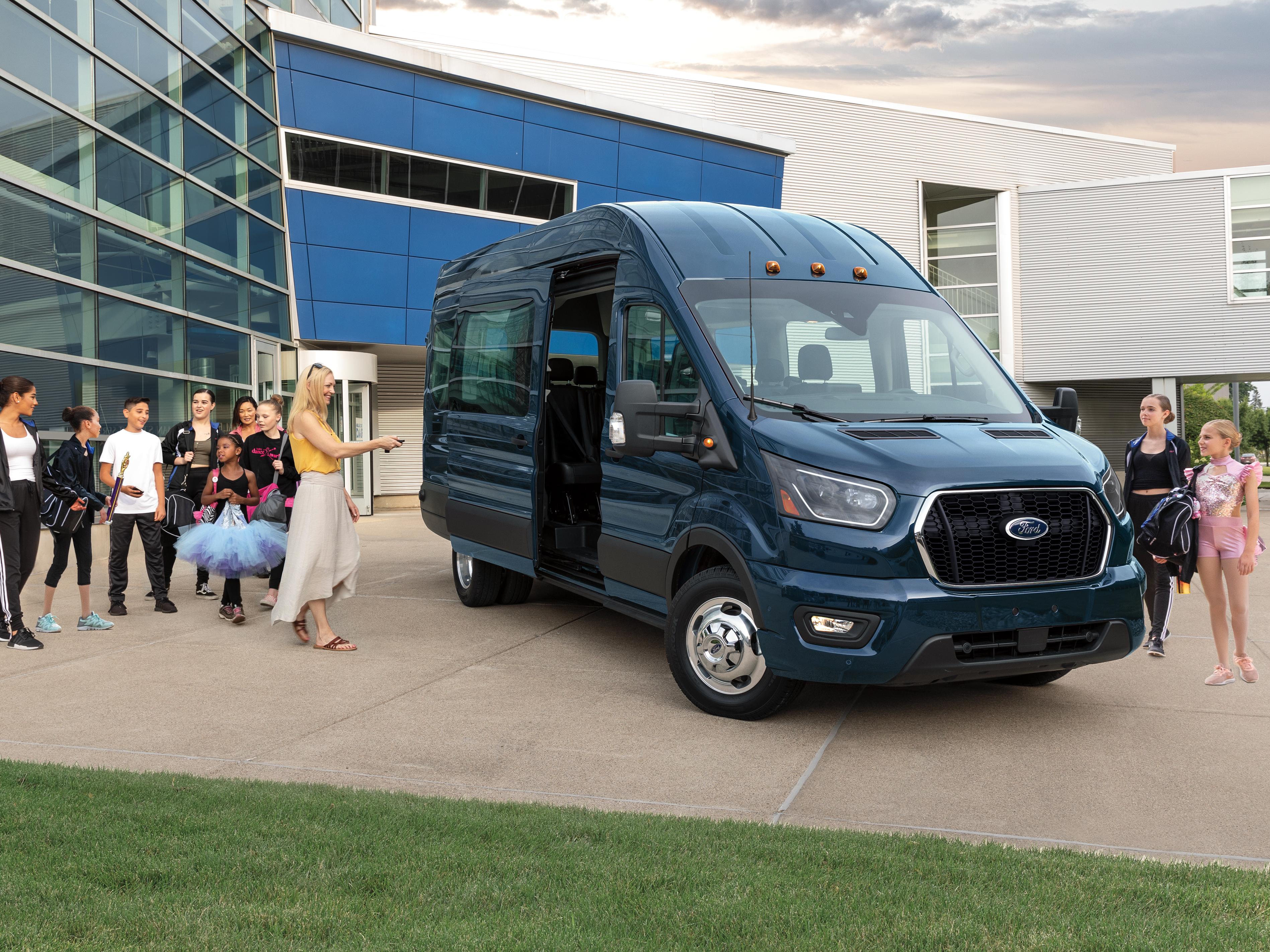 A 2026 Ford Transit® Passenger Van model parked in front of a building with people exiting through the side door