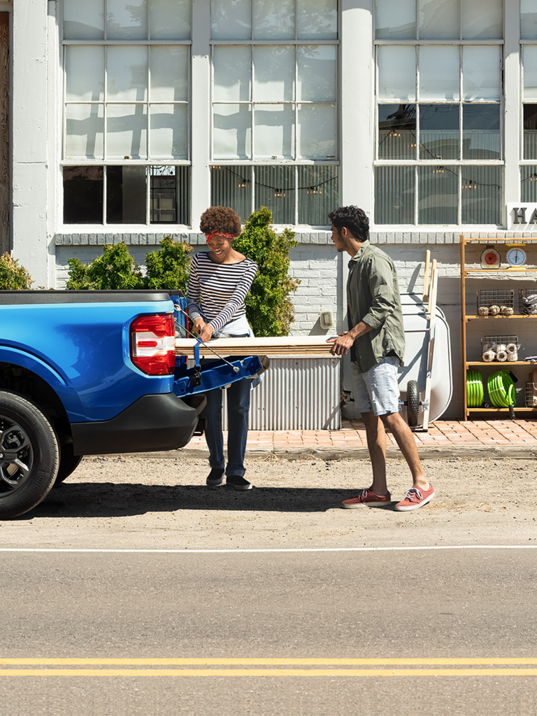 Two people unload plywood from the bed of a Ford Maverick parked in front of a business.
