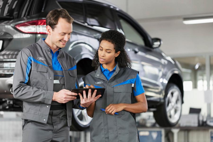 Two Ford employees in front of a vehicle on a car lift looking at a tablet.
