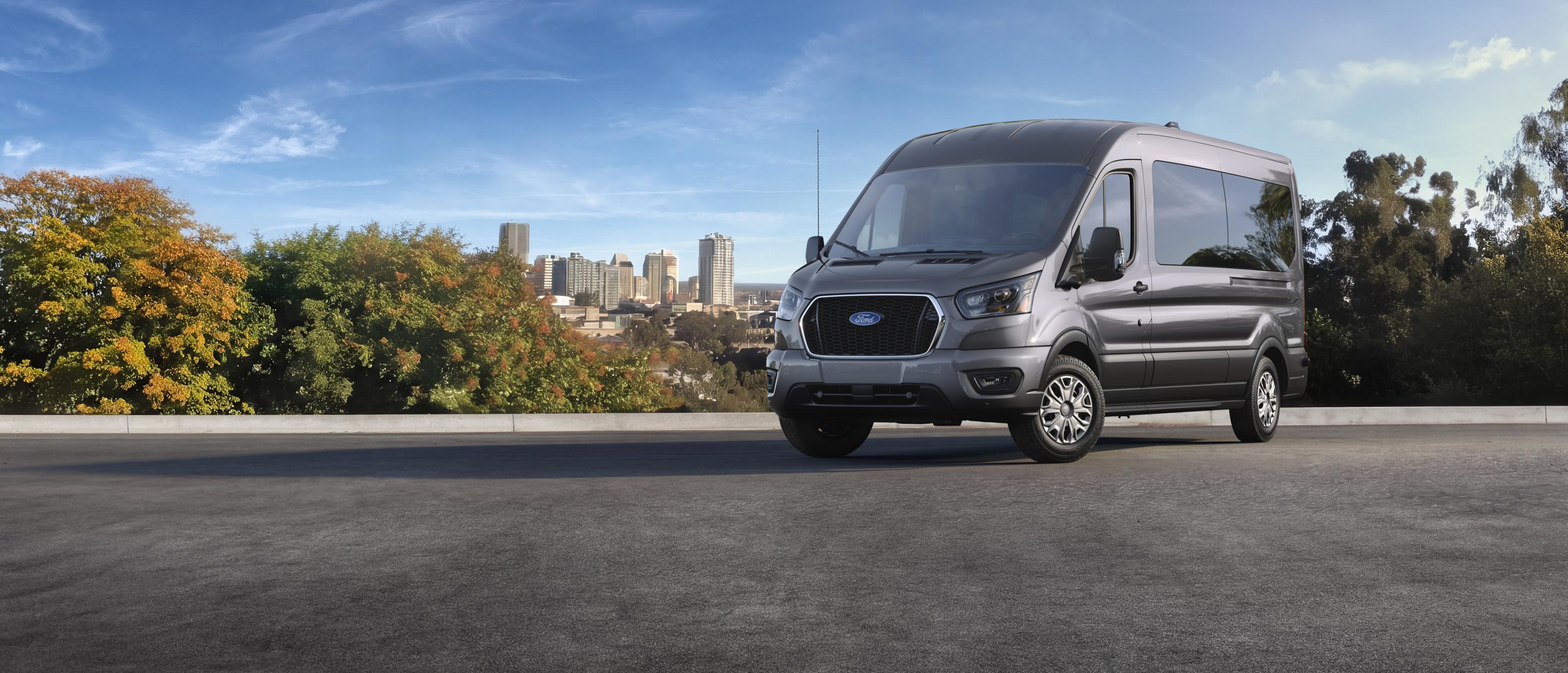 A grey 2027 Ford Transit® Passenger Van parked on an asphalt road with a city skyline visible in the background through trees