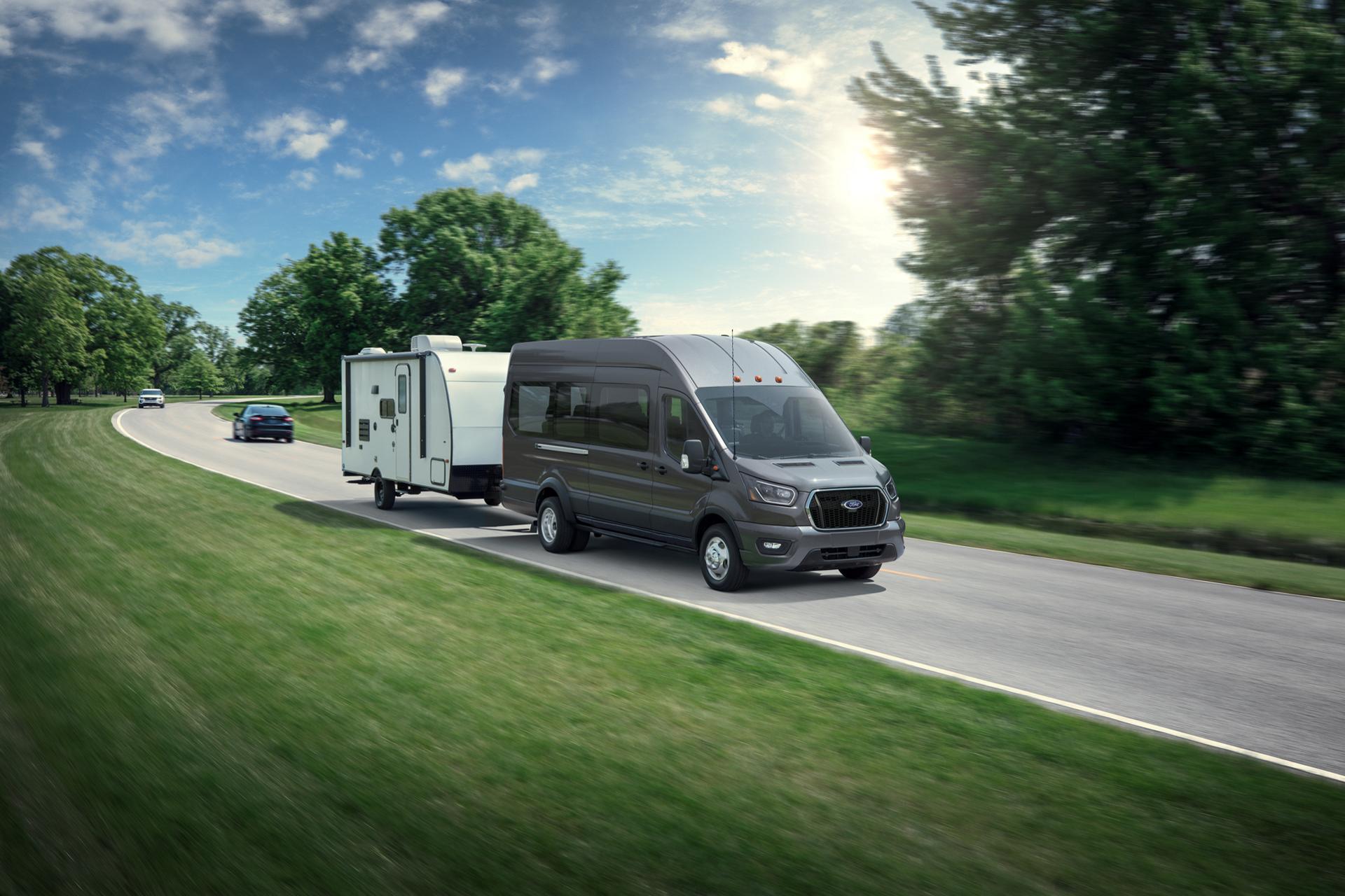 White 2027 Ford Transit® Van towing a small trailer on a winding road with trees