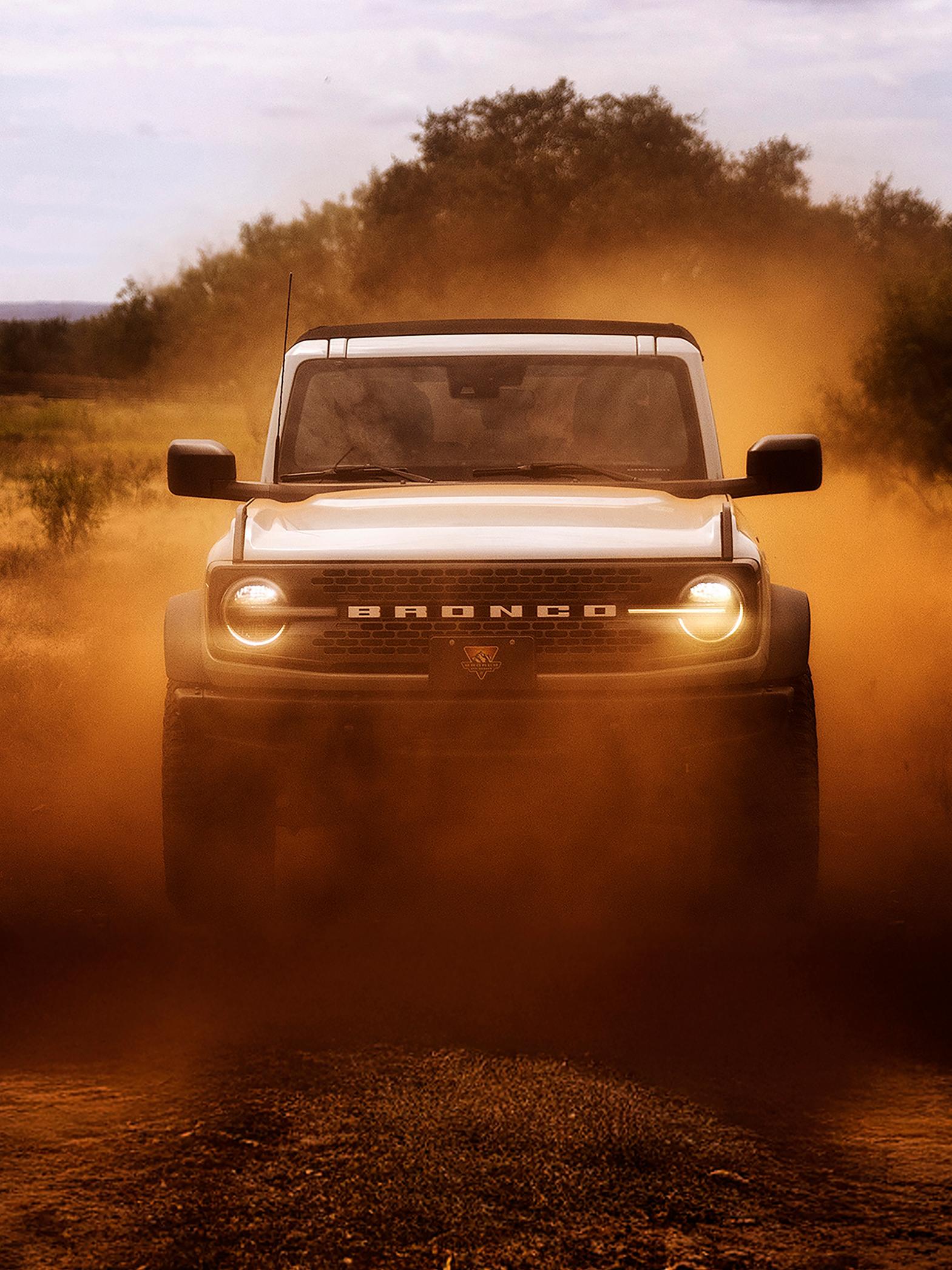 The front of a 2026 Ford Bronco® SUV, its headlights shining through a cloud of red dust
