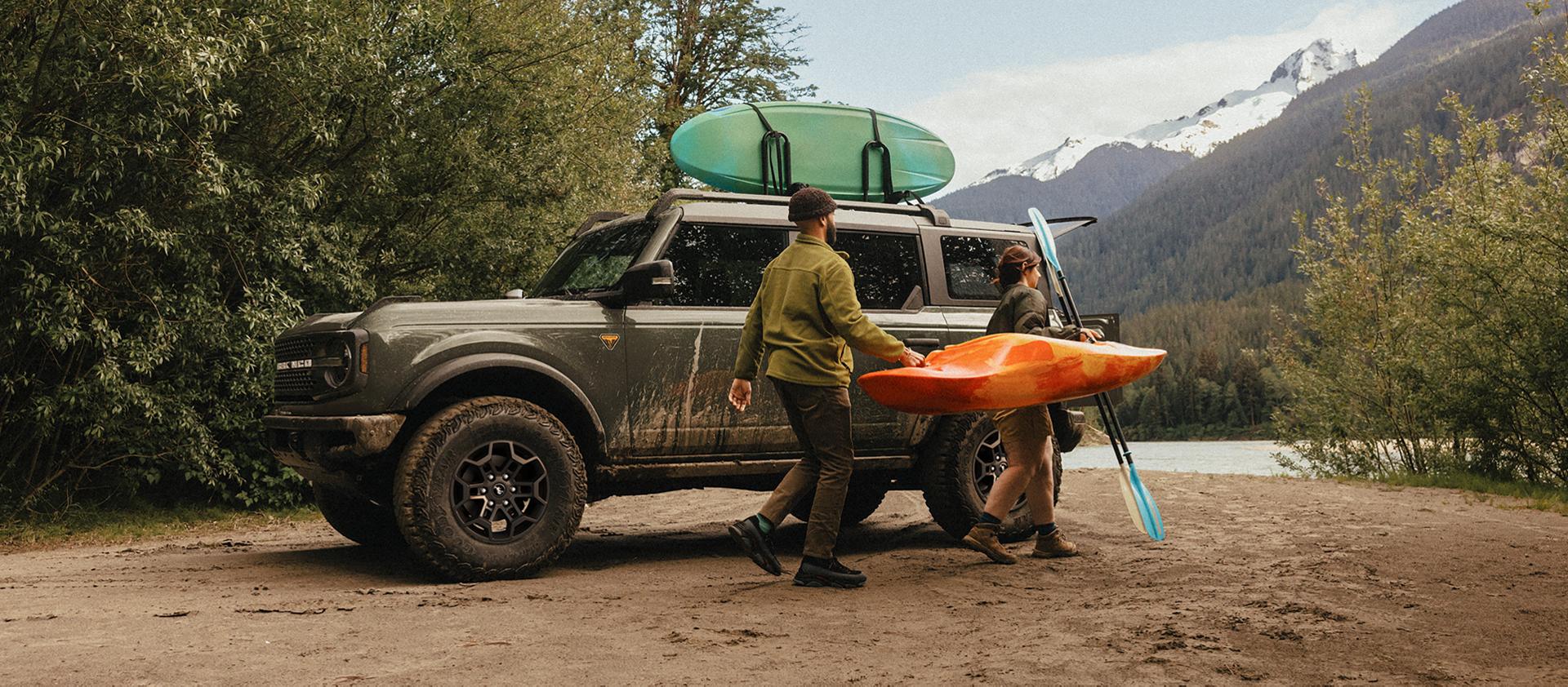 A man and woman carrying a kayak and paddle away from a 2026 Ford Bronco® SUV parked on a mud flat