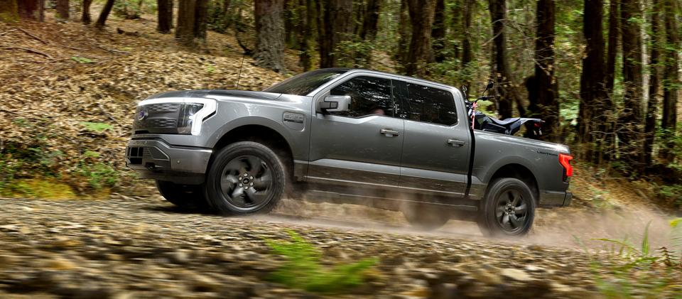 A 2025 Ford F-150® Lightning® pickup driving on a dusty road through the forest with an ATV secured in the cargo bed