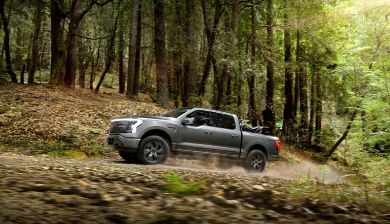 A 2025 Ford F-150® Lightning® pickup driving on a dusty road through the forest with an ATV secured in the cargo bed