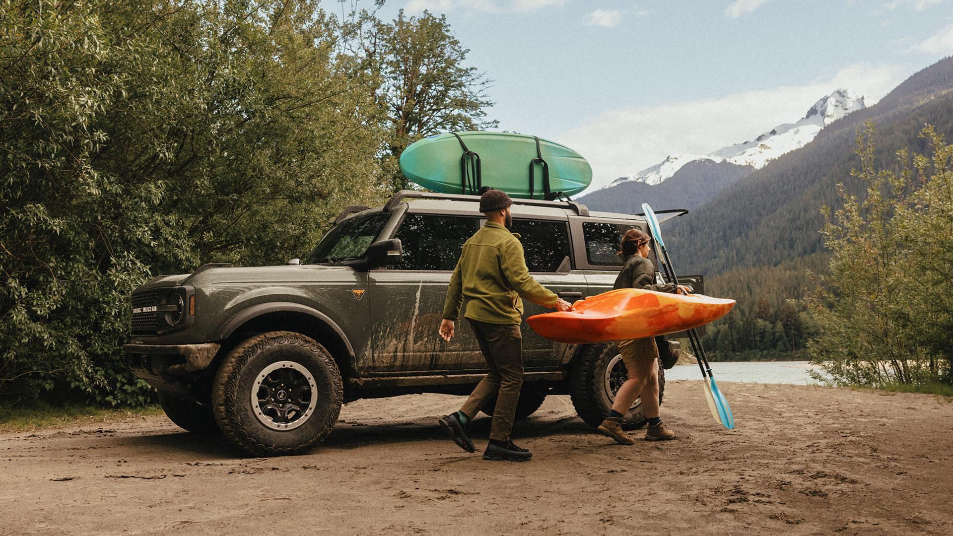 A man and woman carrying a kayak and paddle away from a 2026 Ford Bronco® SUV parked on a mud flat