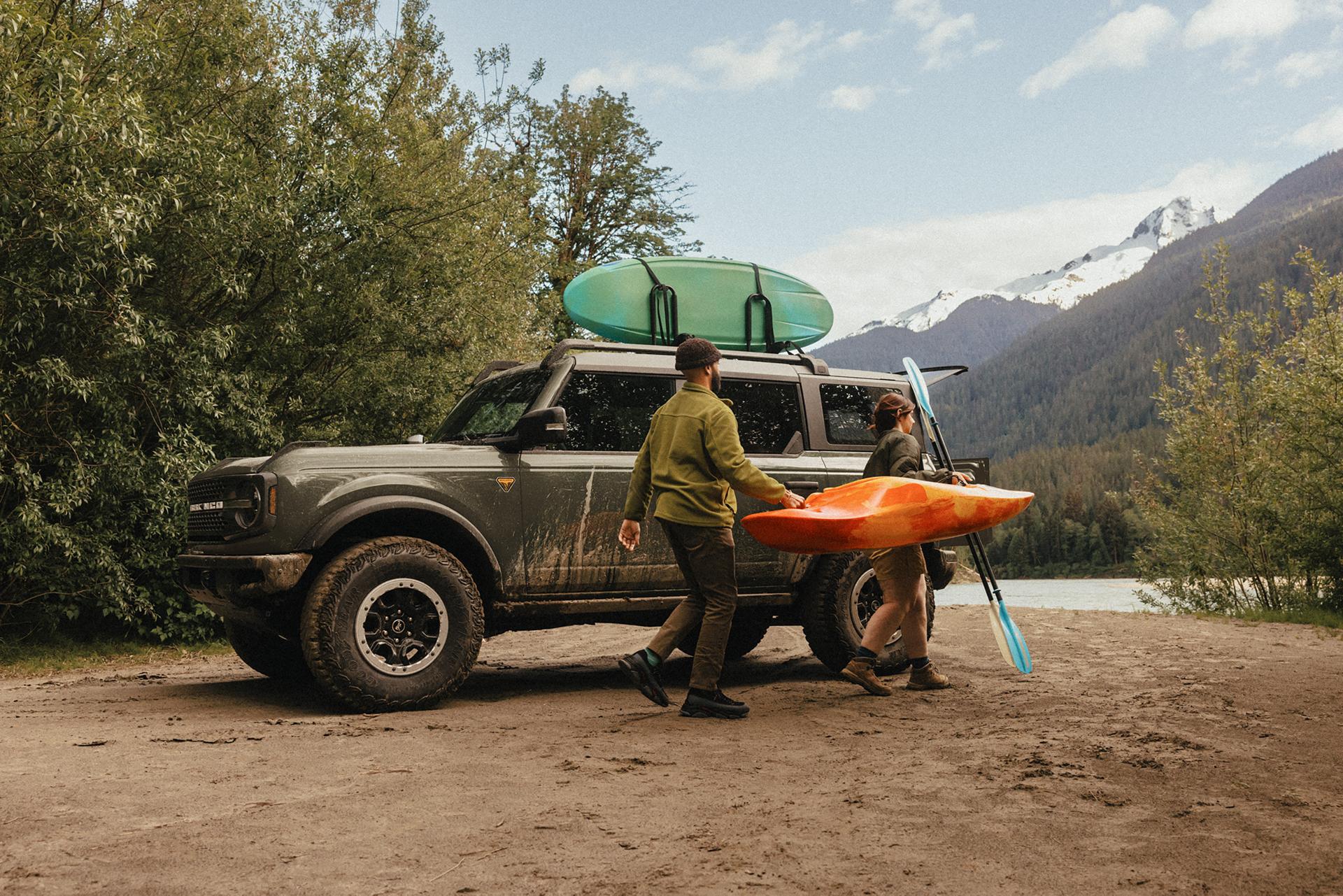 A man and woman carrying a kayak and paddle away from a 2026 Ford Bronco® SUV parked on a mud flat