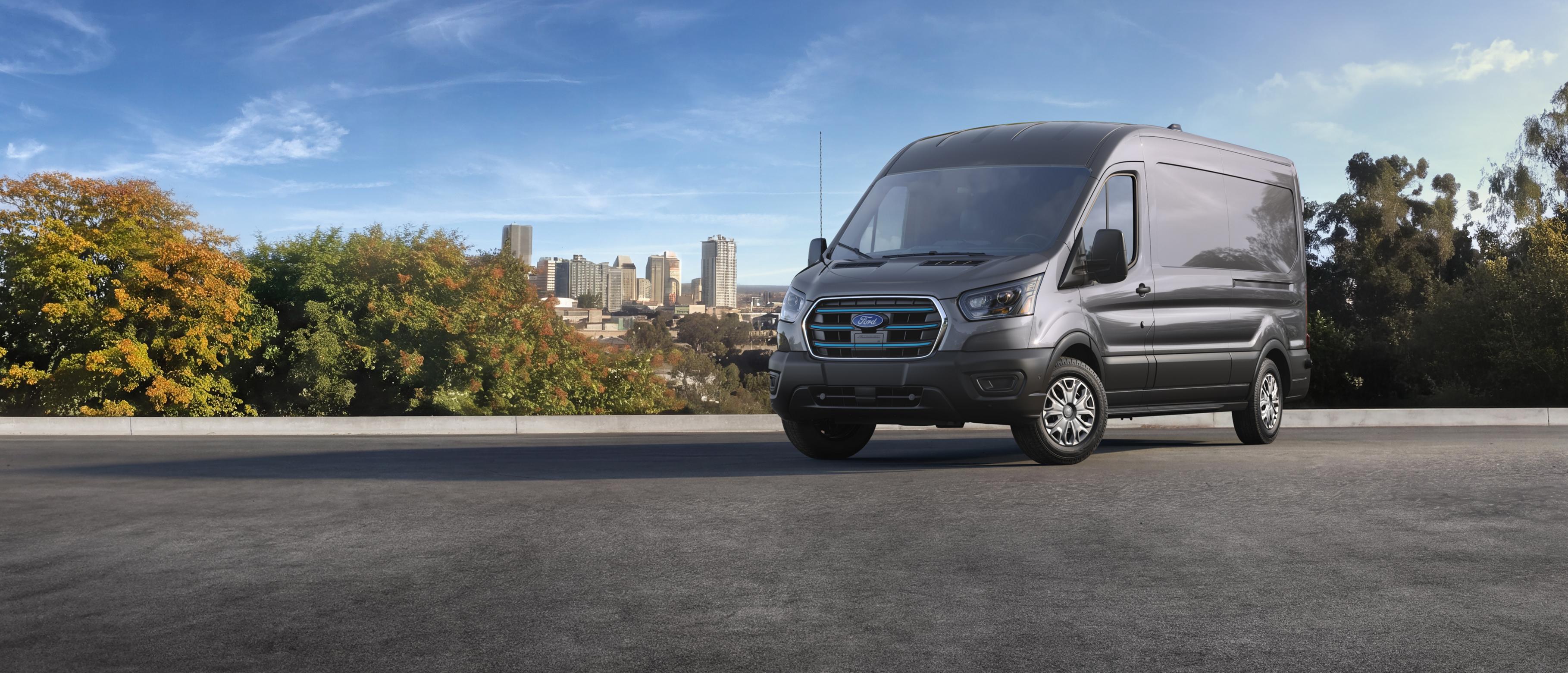 A grey 2027 Ford E-Transit™ Cargo Van parked on an asphalt road, with a city skyline and trees in the background under a clear sky