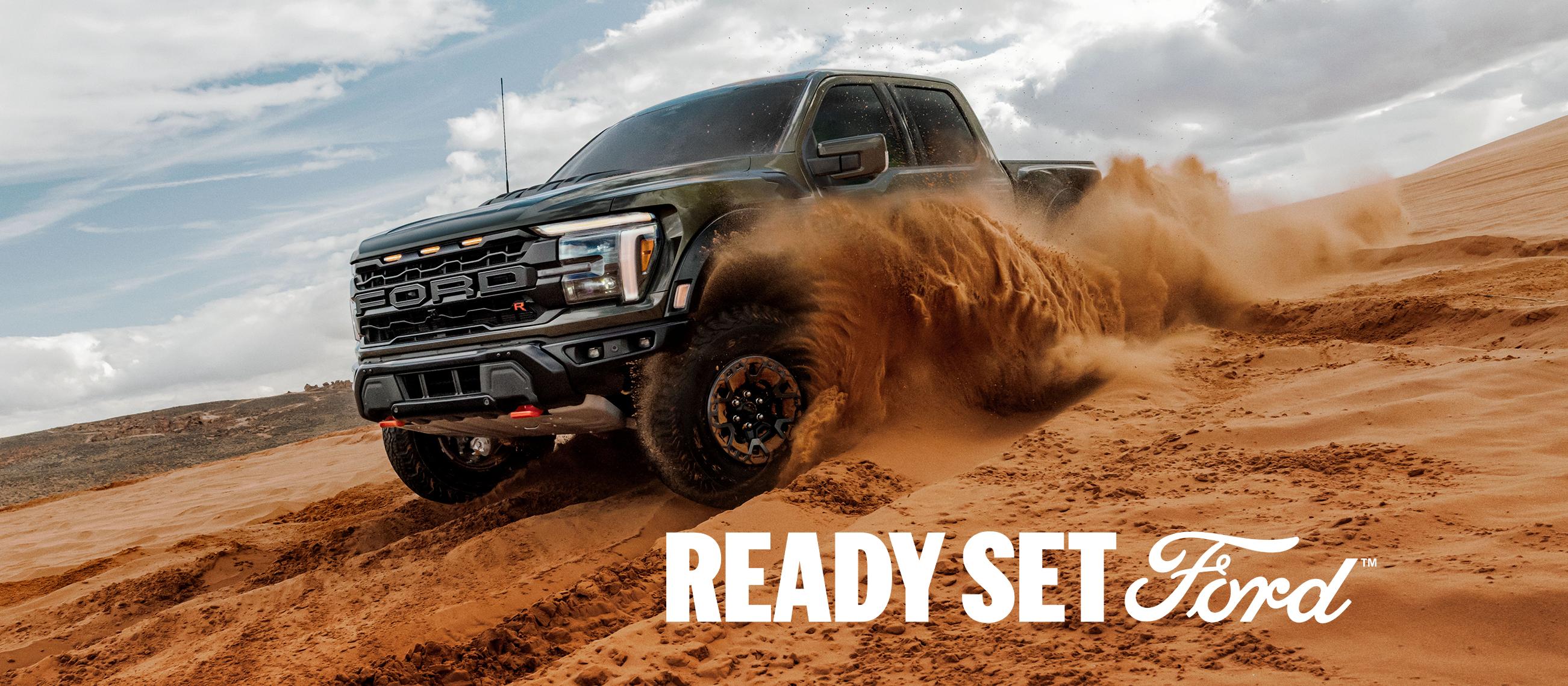 Three-quarter front view of a dark green Ford F-150 Raptor driving off-road through deep sand. White text “Ready Set Ford” appears in the lower left-hand corner. 
