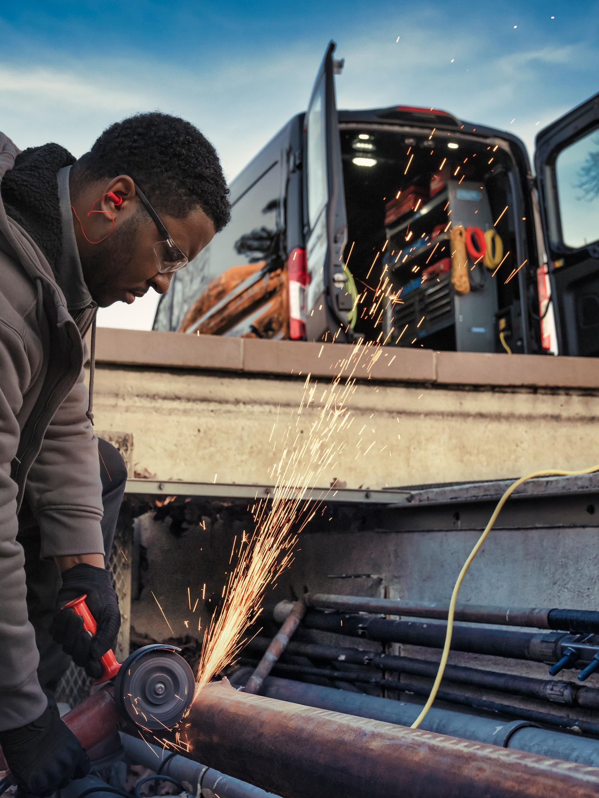 Worker using a grinder, sparks flying, with an open 2027 Ford Transit® Van in the background