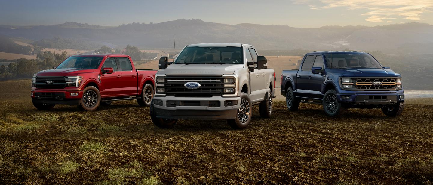 Three Ford F-Series trucks in red, white, and blue parked side by side in a field with rolling hills in the background.