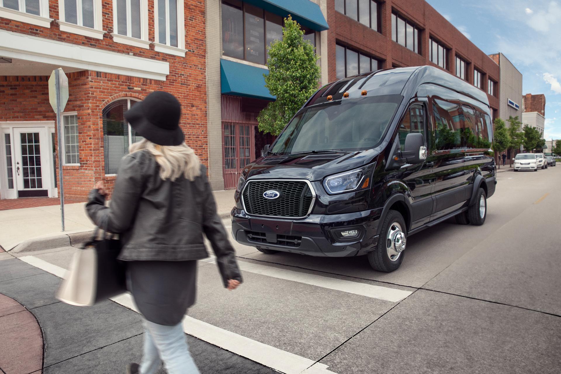 Woman walking in front of a black 2027 Ford Transit® Van parked on a city street with brick buildings